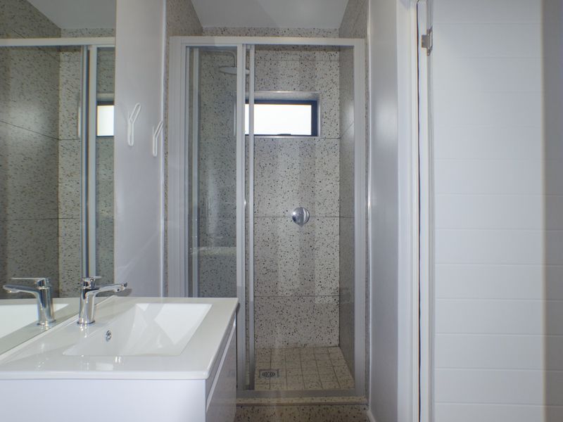 A clean, modern bathroom featuring a glass-enclosed shower, a white vanity basin with a mixer tap, and neutral speckled wall tiling.
