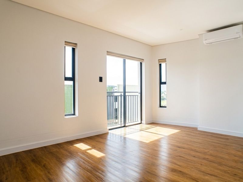 A bright, empty bedroom featuring modern laminate flooring, a wall-mounted air conditioning unit, and access to a private balcony through sliding glass doors.