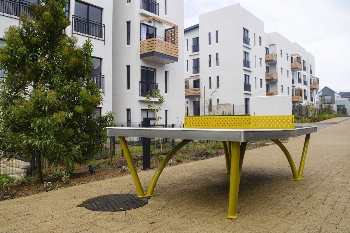 Outdoor communal area featuring a paved walkway, landscaped garden beds, and a fixed table tennis table set against modern apartment buildings.