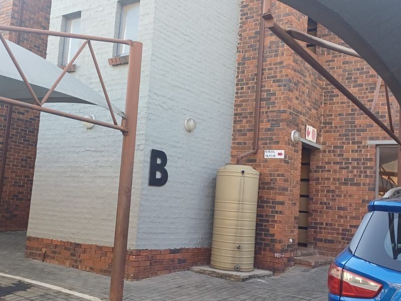 Exterior view of a townhouse complex entrance featuring brick facade, paved parking area with shade cloth carports, and a communal water storage tank.