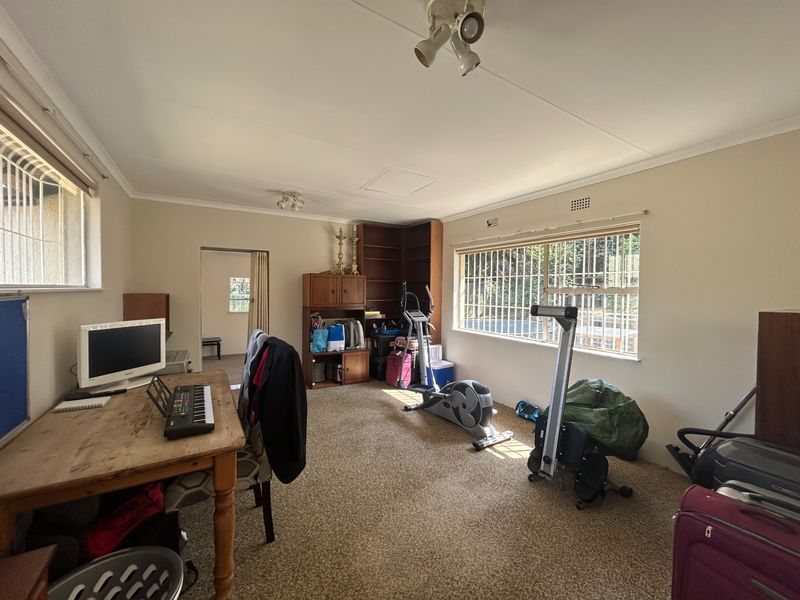 A spacious living area featuring carpeted flooring, built-in shelving, and large windows fitted with security bars, offering a versatile space for various uses.
