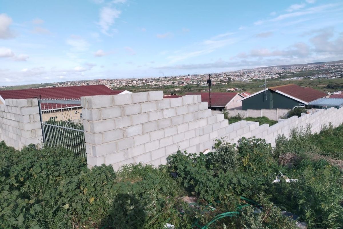 A view of a residential plot featuring an unfinished concrete block boundary wall and metal gate, overlooking the surrounding neighborhood.