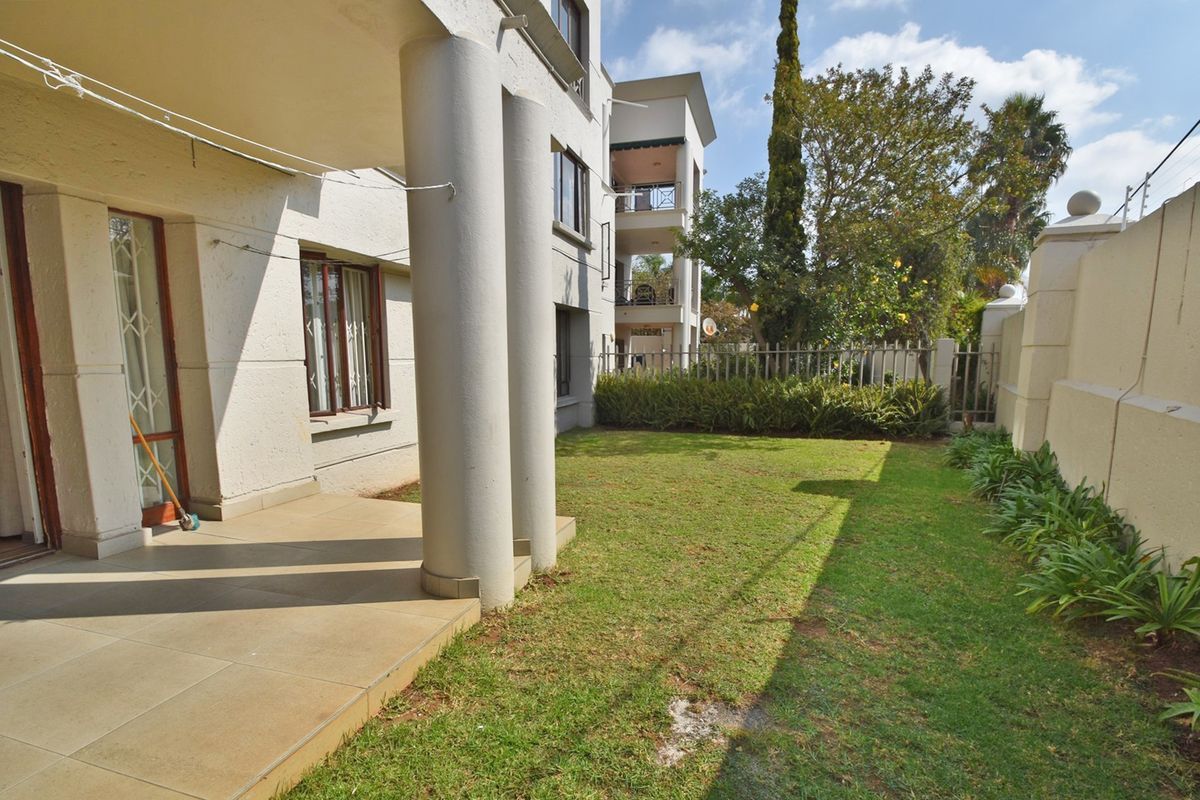 A private rear garden featuring a tiled patio area with structural columns, enclosed by a perimeter wall and metal security fencing.