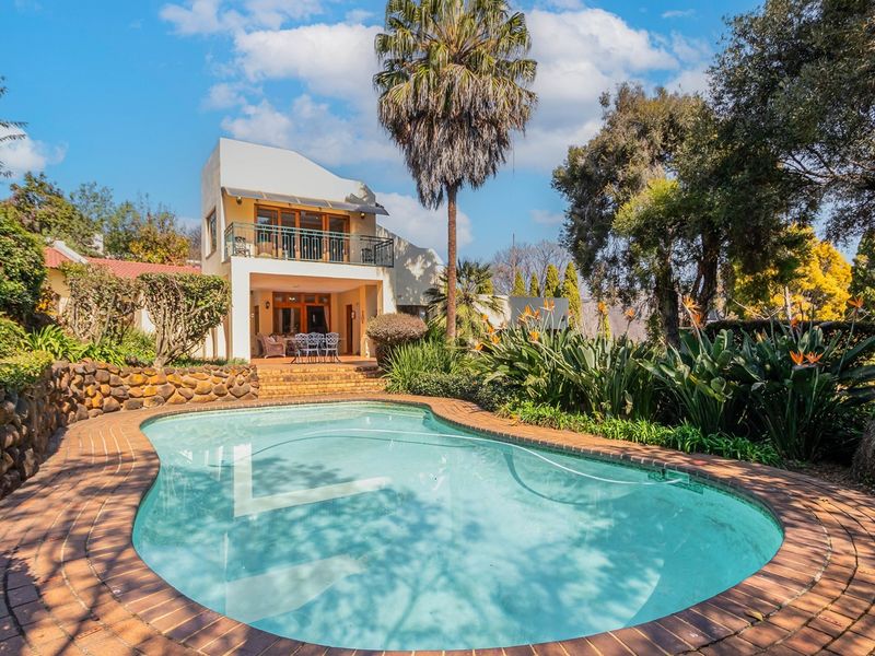 A well-maintained swimming pool with brick coping, set in a landscaped garden with a stone retaining wall and a two-story home with a balcony.