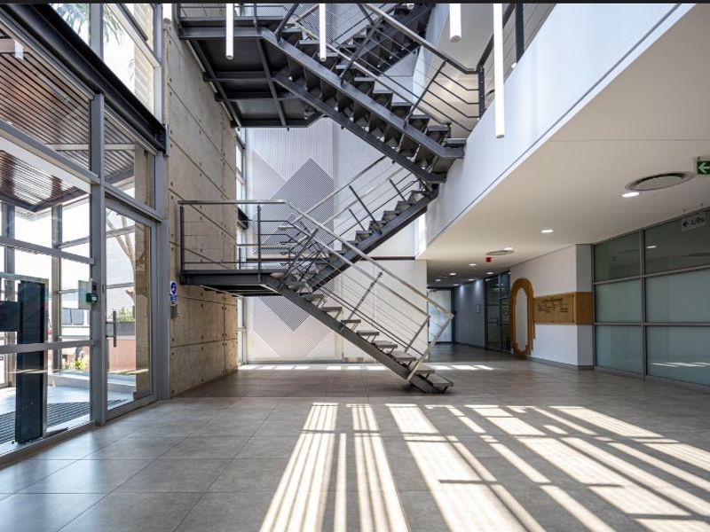 Modern office building entrance lobby featuring a striking industrial steel staircase, exposed concrete wall, and expansive glass facade with natural light.