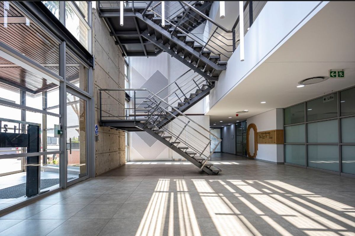 Modern office building entrance lobby featuring a striking industrial steel staircase, exposed concrete wall, and expansive glass facade with natural light.