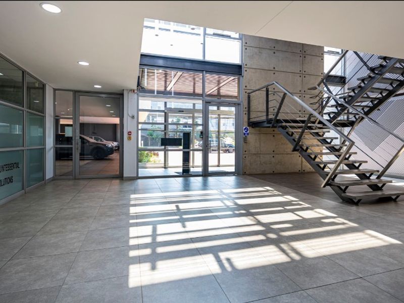 Modern office building entrance lobby featuring high ceilings, exposed concrete architectural elements, a steel staircase, and large glass partitions.