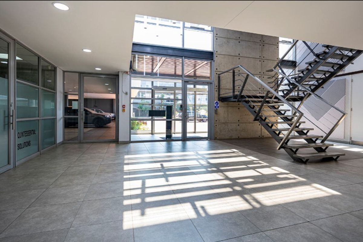 Modern office building entrance lobby featuring high ceilings, exposed concrete architectural elements, a steel staircase, and large glass partitions.