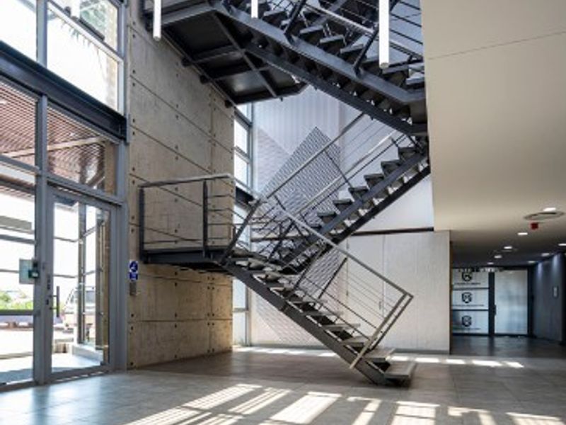 Modern office building entrance featuring a striking industrial steel staircase, exposed concrete walls, and expansive glass windows providing abundant natural light.