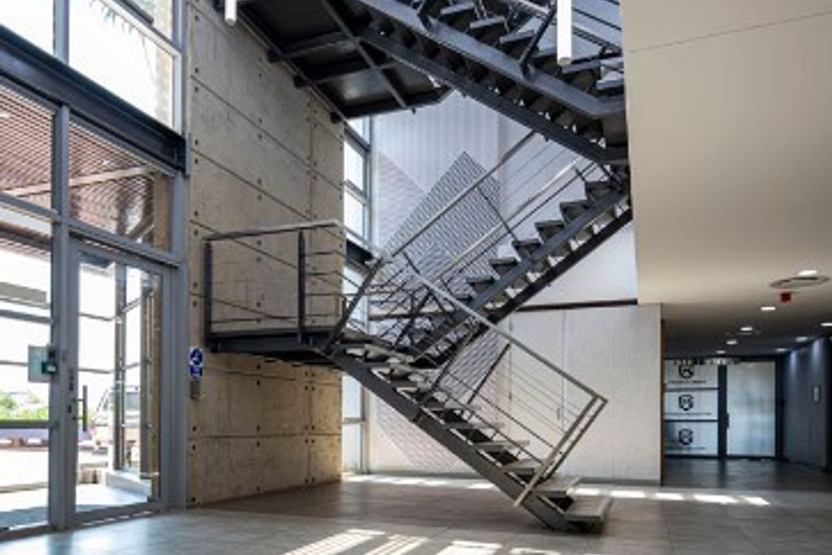 Modern office building entrance featuring a striking industrial steel staircase, exposed concrete walls, and expansive glass windows providing abundant natural light.
