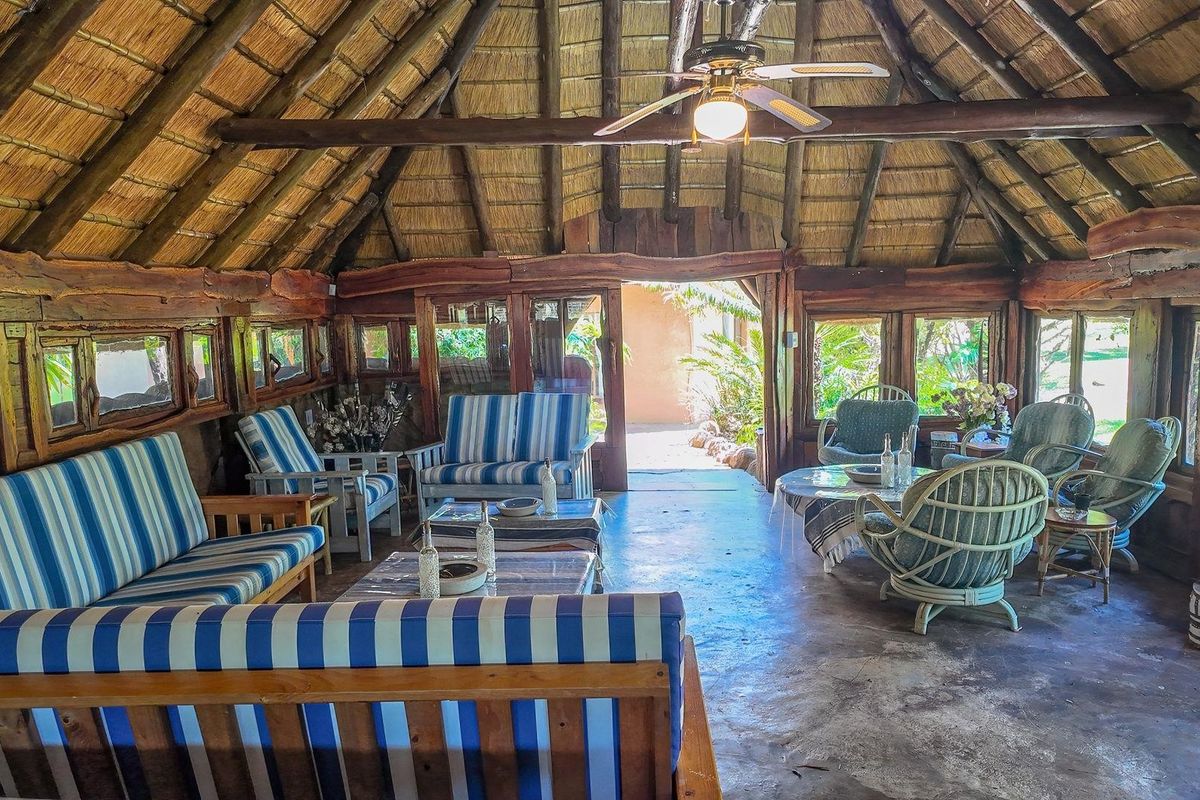 Rustic living area featuring a traditional thatched roof, exposed timber beams, ceiling fan, and concrete flooring with direct access to the garden.