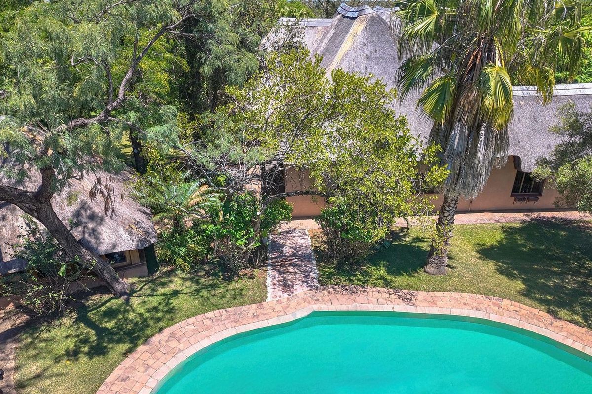 Aerial view of a farm property featuring a swimming pool, brick-paved pathways, and traditional thatched-roof buildings set within a lush, tree-filled garden.