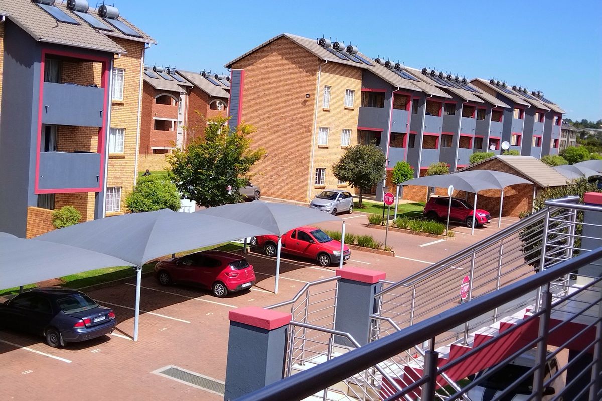 An elevated view of an apartment complex courtyard featuring brick buildings, solar water heating systems, and covered parking bays with shade cloth structures.