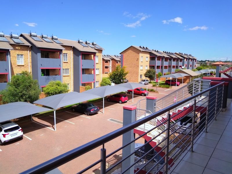 Elevated view of the apartment complex exterior, featuring paved parking with shade cloth carports, solar water heaters, and a tiled balcony with steel railings.