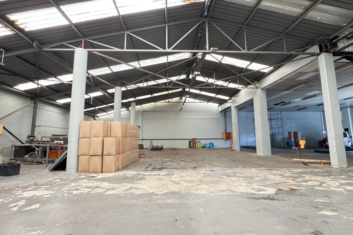 Spacious industrial warehouse featuring high exposed steel truss ceilings, natural light from skylights, concrete flooring, and structural support columns.
