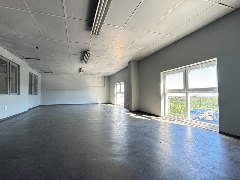 A spacious, empty industrial office area featuring a suspended ceiling, fluorescent lighting, and large windows providing natural light.