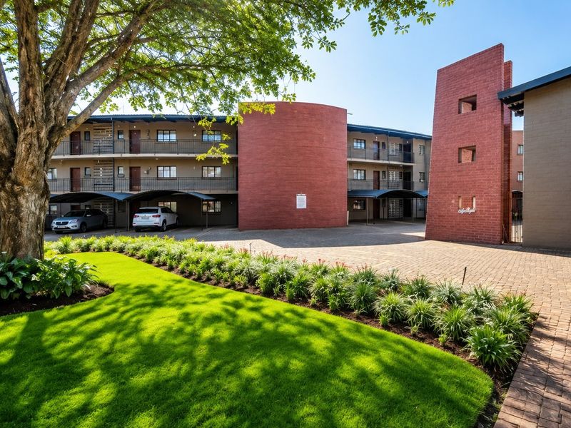A well-maintained apartment complex exterior featuring a paved driveway, covered parking bays, and landscaped garden areas with mature trees.