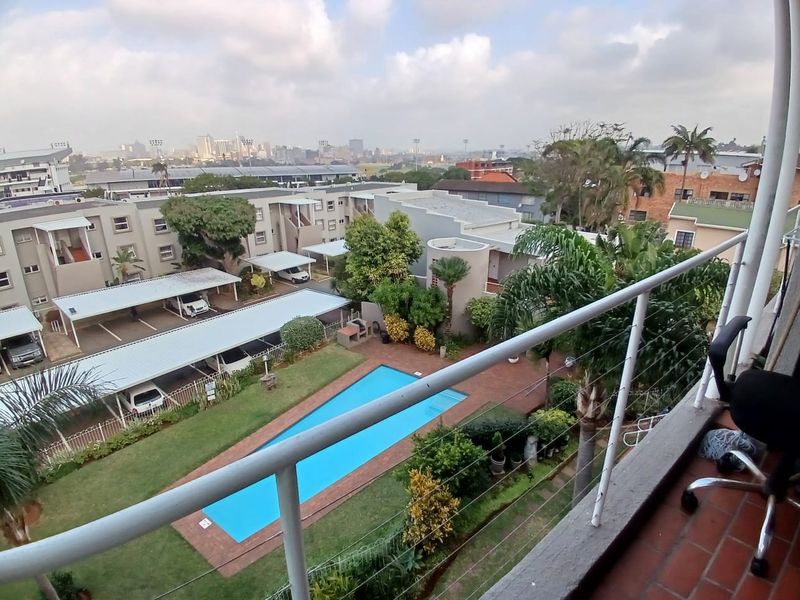 Elevated balcony view overlooking the apartment complex grounds, featuring a swimming pool, landscaped garden, and covered parking bays.