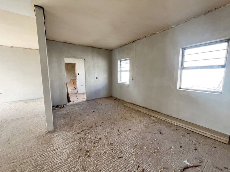 An unfinished interior living space featuring plastered walls, steel-framed windows, and a structural column, currently in a shell condition awaiting final finishes.