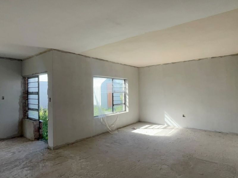 An empty, unfinished living area featuring plastered walls, aluminium window frames, and a glass door, awaiting final flooring and decorative finishes.