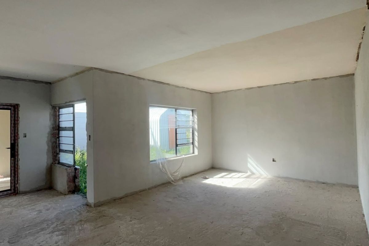 An empty, unfinished living area featuring plastered walls, aluminium window frames, and a glass door, awaiting final flooring and decorative finishes.