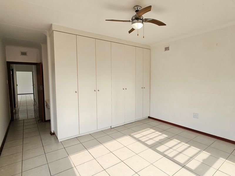 A bright, empty bedroom featuring neutral tiled flooring, a ceiling fan, and extensive built-in cupboard storage.