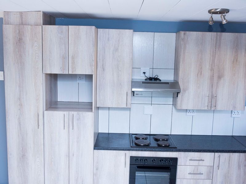 A functional kitchen featuring light wood-grain cabinetry, dark laminate countertops, a built-in oven, electric hob, and a tiled backsplash.