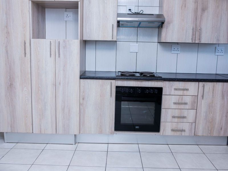 A clean, modern kitchen featuring light wood-grain cabinetry, dark countertops, a built-in oven, electric hob, and an extractor fan with tiled flooring.
