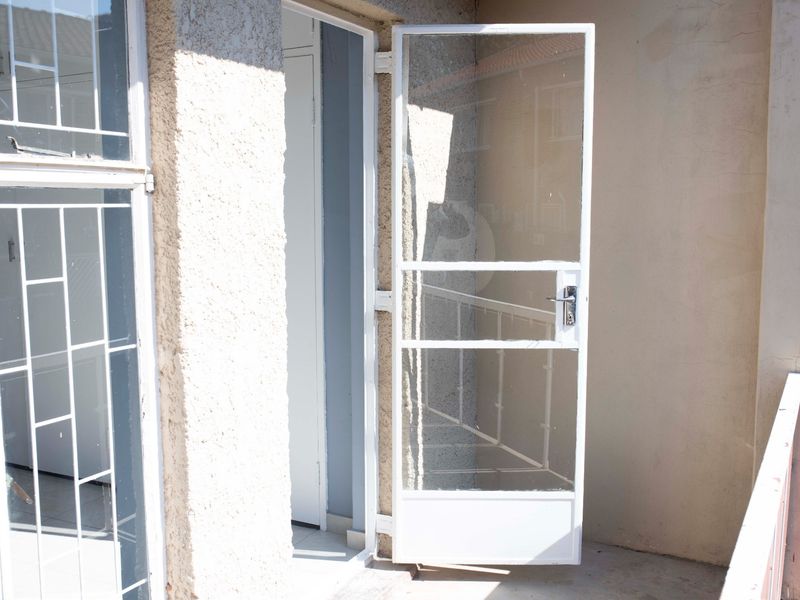 A small balcony area featuring a white security gate and metal balustrade, showing signs of wear on the exterior wall finish.