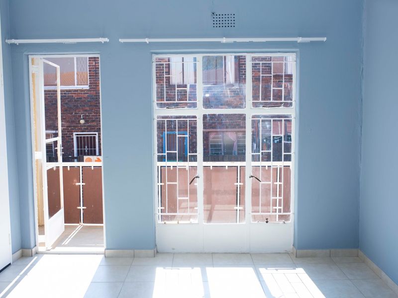 An empty living room featuring tiled flooring, blue walls, and direct access to a small balcony through a security gate and steel-framed windows.