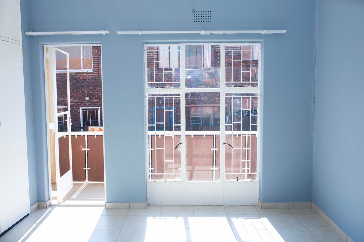An empty living room featuring tiled flooring, blue walls, and direct access to a small balcony through a security gate and steel-framed windows.