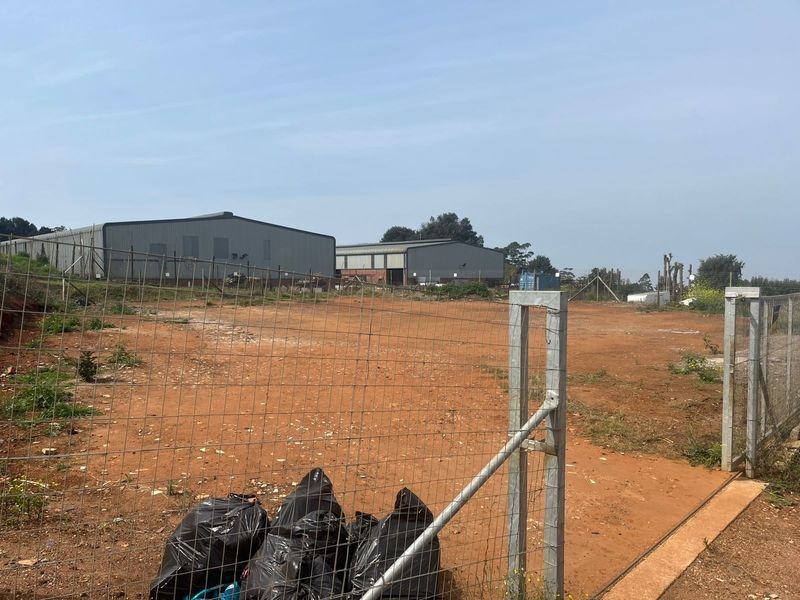 A view of a vacant agricultural plot featuring wire mesh fencing, a metal gate, and industrial warehouse structures in the background.