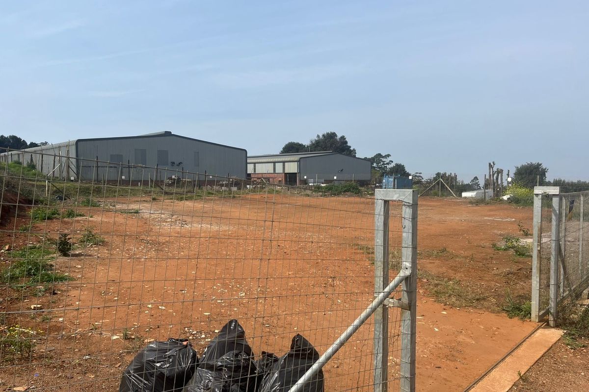 A view of a vacant agricultural plot featuring wire mesh fencing, a metal gate, and industrial warehouse structures in the background.