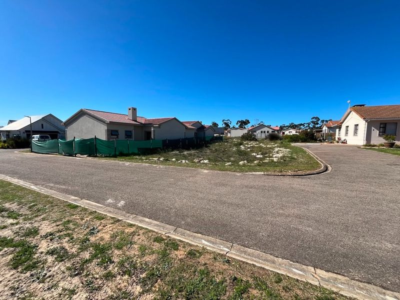A street-level view of a vacant residential plot situated within a developed neighborhood, featuring paved road access and established surrounding homes.