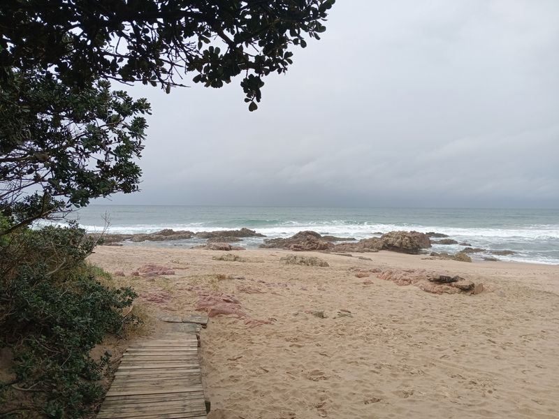 A scenic view of the coastline featuring a wooden boardwalk leading onto a sandy beach with rocky outcrops and ocean waves.