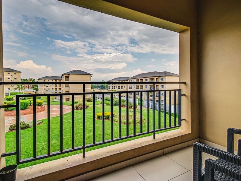 A covered balcony featuring a metal balustrade and tiled flooring, offering an elevated view over the apartment complex grounds and landscaped gardens.