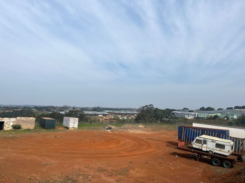 An elevated view of a vacant agricultural plot featuring unpaved ground and perimeter fencing with surrounding industrial buildings in the distance.