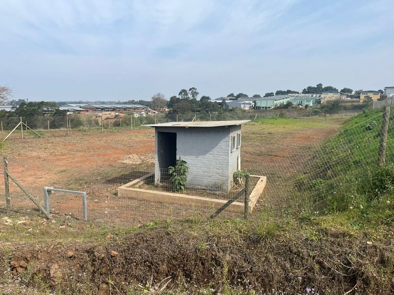 A view of a vacant agricultural plot featuring a small utility structure, enclosed by wire mesh fencing with surrounding landscape.