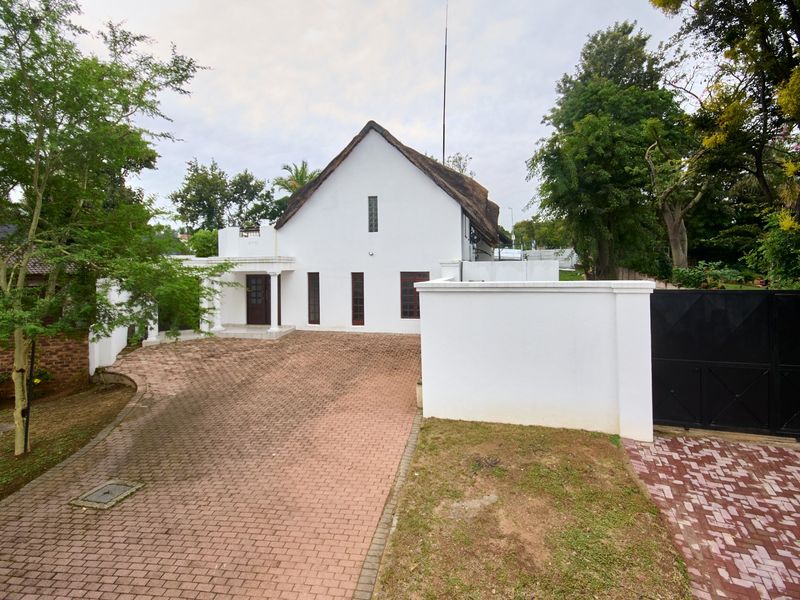 Front exterior view of a white house with a thatched roof, featuring a paved driveway, boundary wall, and gated entrance.