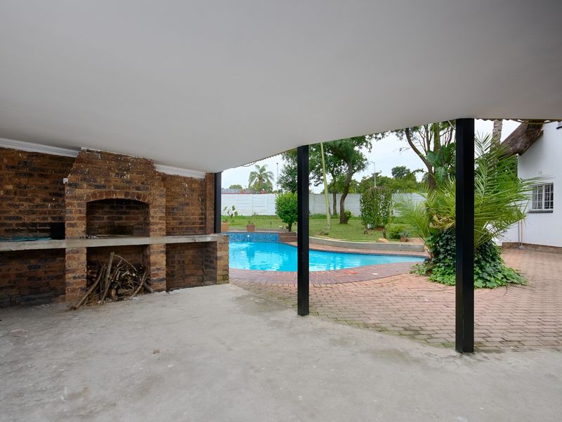 Covered outdoor patio featuring a built-in brick braai, overlooking a swimming pool and garden area with brick paving.