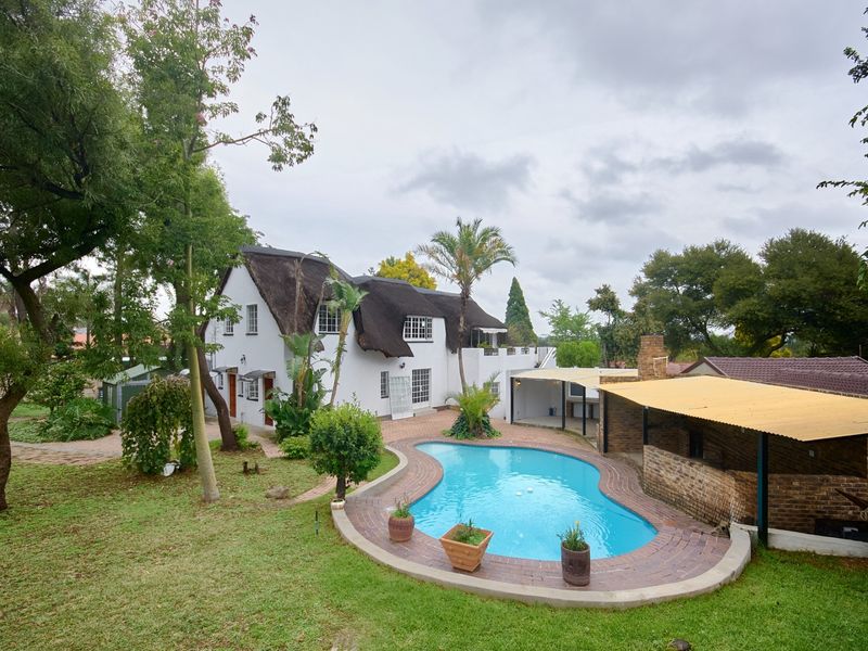 Rear view of a property featuring a swimming pool, brick patio, covered entertainment area, and a distinctive thatched roof house set in a garden.