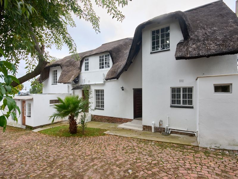 A charming double-story home featuring a traditional thatched roof, white stucco exterior, and a classic brick-paved driveway entrance.