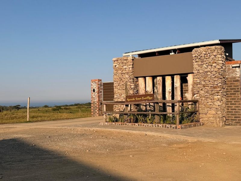 The entrance facade of Gqunube Green Ecovillage featuring natural stone cladding, timber posts, and a paved driveway approach.