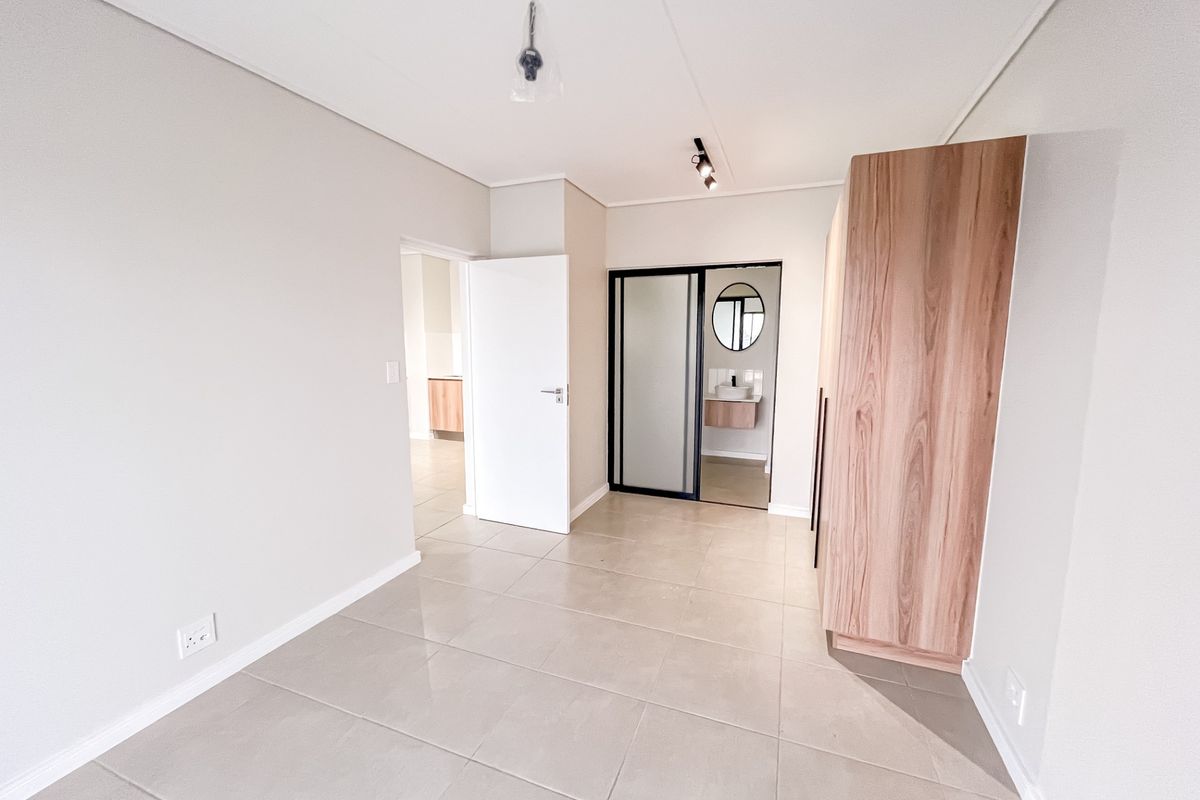 A bright, modern bedroom featuring neutral tiled flooring, built-in wooden cabinetry, and a sliding door leading to an en-suite bathroom.