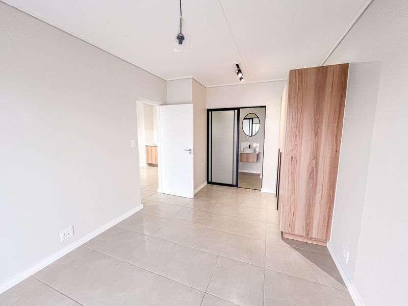 A bright, modern bedroom featuring neutral tiled flooring, built-in wooden cabinetry, and a sliding door leading to an en-suite bathroom.