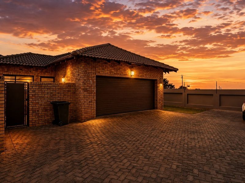 A well-maintained townhouse exterior featuring a brick facade, tiled roof, automated garage door, and paved driveway at sunset.