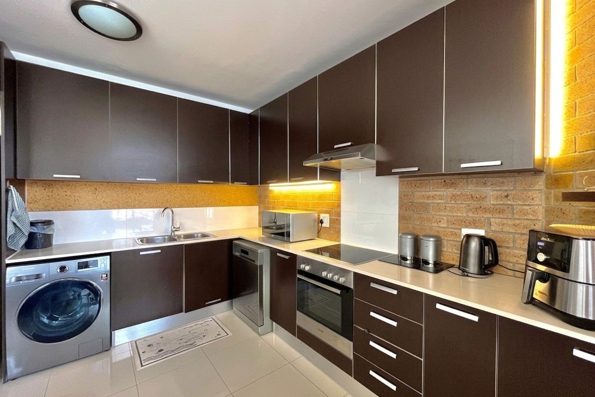 Modern kitchen featuring dark cabinetry, stainless steel appliances, integrated oven, electric cooktop, and a stylish exposed brick backsplash with under-cabinet lighting.