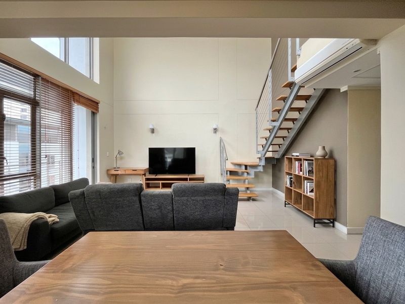 Spacious open-plan living area featuring double-volume ceilings, a modern staircase, large windows for natural light, and neutral tiled flooring.