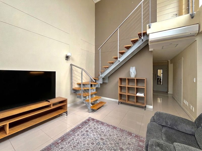 Spacious living area featuring a modern staircase with metal balustrades, high ceilings, tiled flooring, and a wall-mounted air conditioning unit.