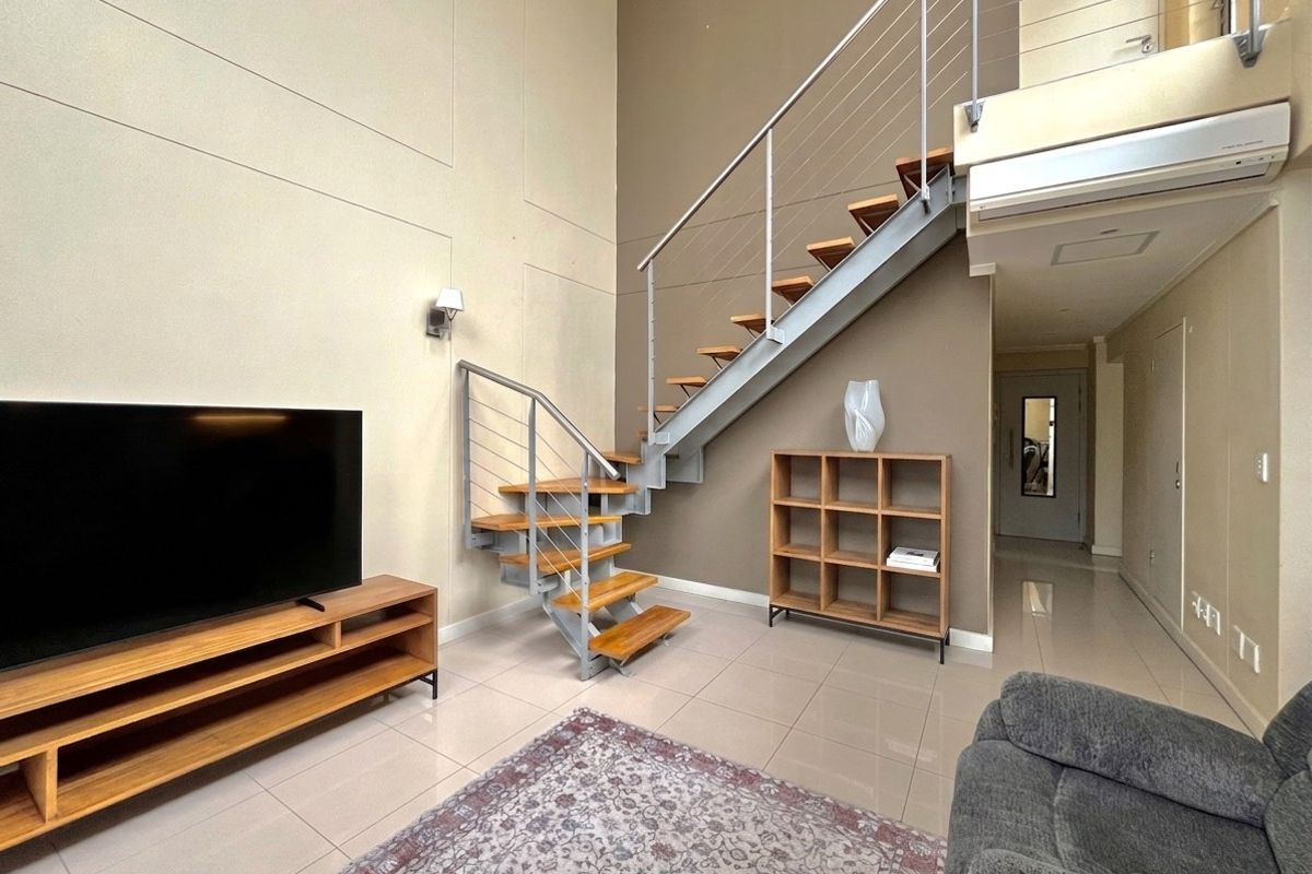 Spacious living area featuring a modern staircase with metal balustrades, high ceilings, tiled flooring, and a wall-mounted air conditioning unit.